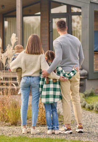 Happy family. View from back of man woman with child and girl standing together hugging and admiring their cozy home on fine autumn day